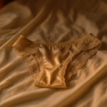 Braguitas de satén color beige con encaje delicado, reposando sobre sábanas claras iluminadas por luz cálida.