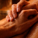 Primer plano cinematográfico y detallado de dos manos entrelazadas con piel húmeda y vibrante bajo una luz cálida y granulada; al fondo, el reflejo ámbar en una jarra de cristal que simboliza la transparencia y la sed del deseo sagrado.